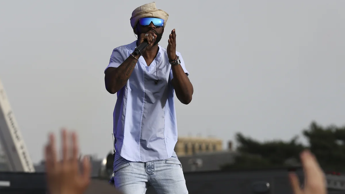 Fally Ipupa chante au micro sur scène devant une foule, bras levés au premier plan