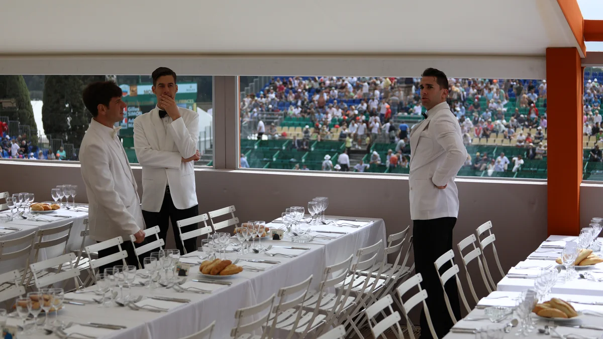 Une loge d'hospitalité avec tables dressées et personnel en veste blanche, donnant sur les tribunes pleines d'un tournoi de tennis
